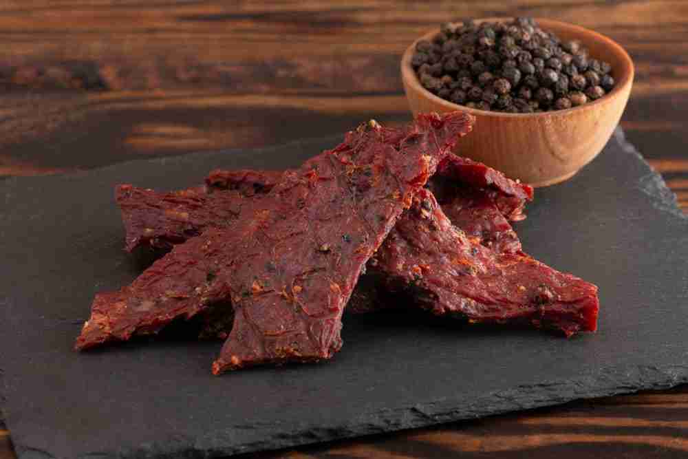 slices of beef jerky presented on a slate with a bowl of peppercorns